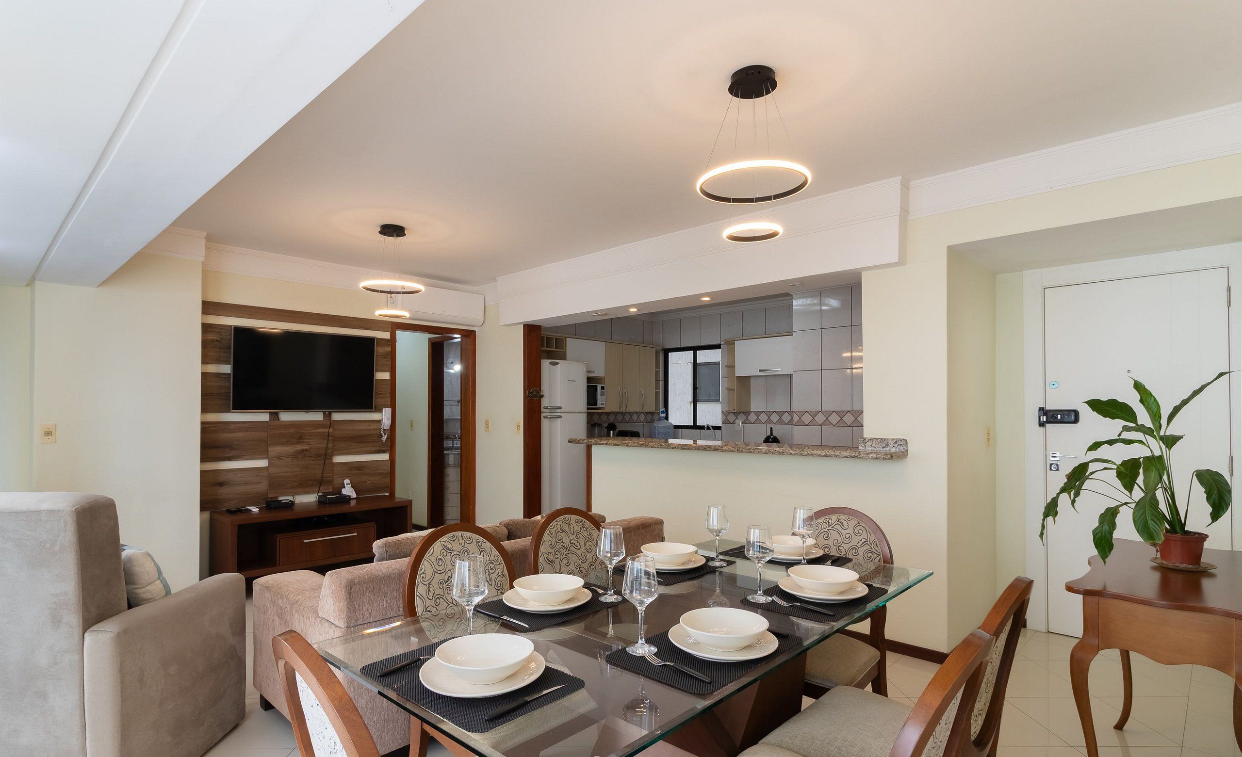A modern dining area with a glass table set for six, surrounded by cushioned chairs. A kitchen is visible in the background, and a potted plant is on a side table.