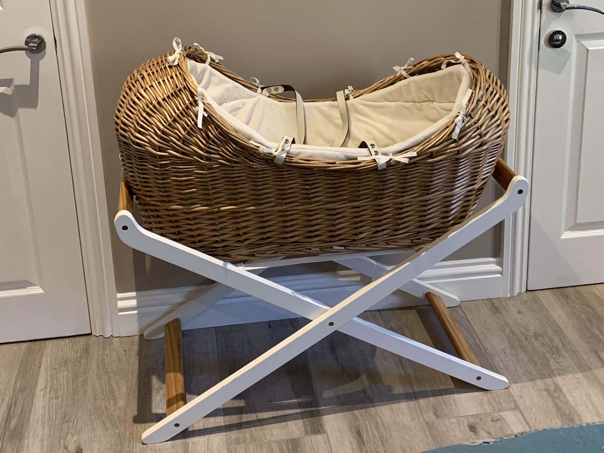 Valkoiseen telineeseen asetettu rottinkinen vauvansänky huoneessa, jossa on puulattia ja valkoiset ovet.