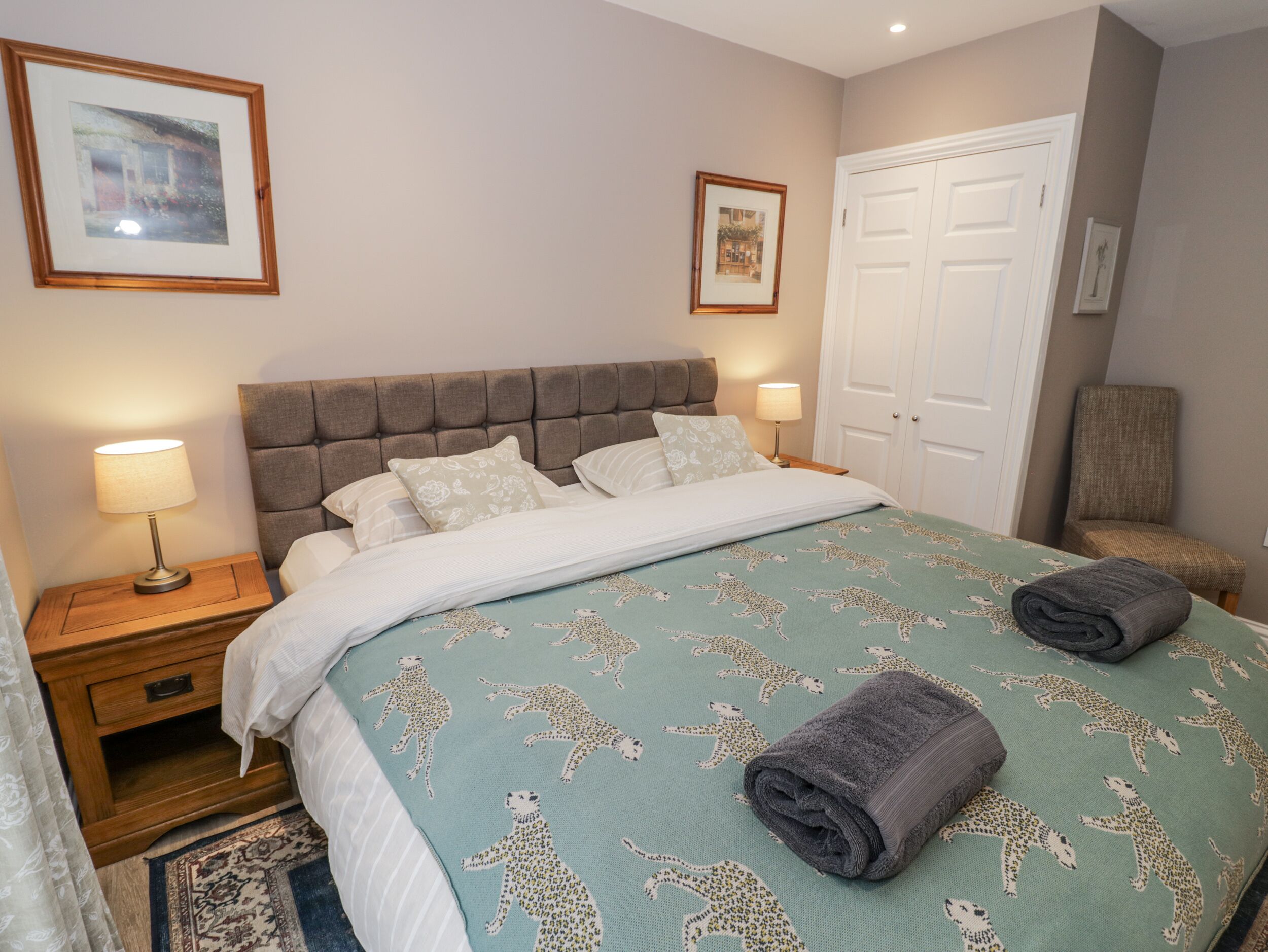 A neatly made bed with a patterned duvet, two bedside lamps, a wooden nightstand, a chair, and a door.