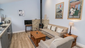 A living room with a sofa, coffee table, TV, fireplace, kitchen area, and decorative wall art. - Rose Stables (Bampton)