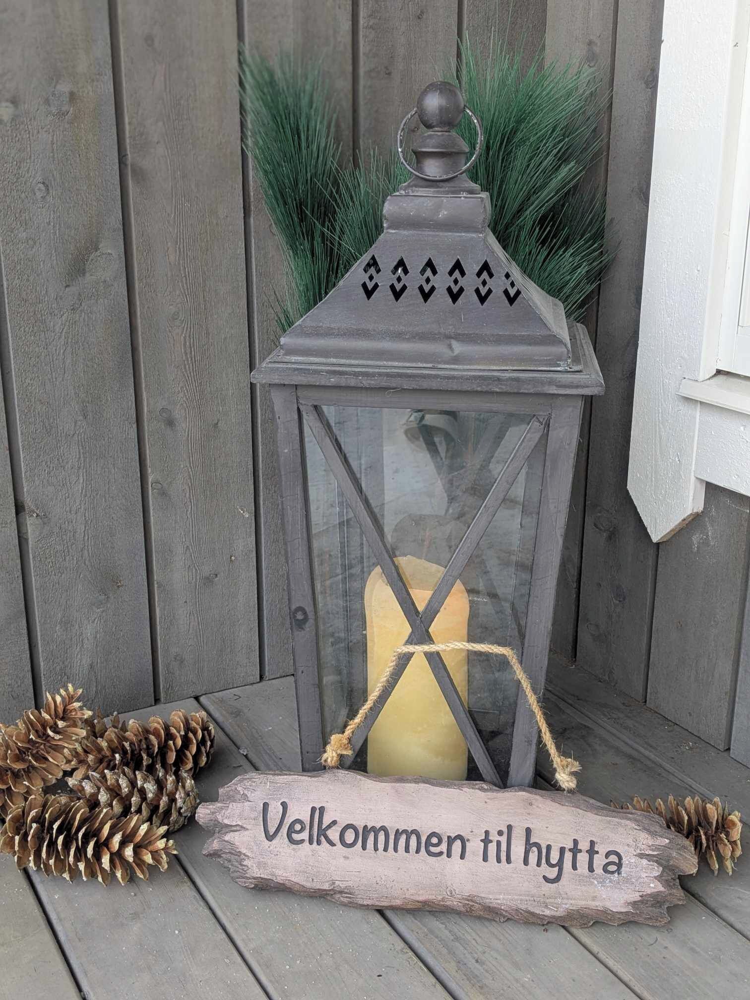 Une lanterne avec une bougie à l’intérieur, posée sur une surface en bois, et sur laquelle est accroché un panneau indiquant : « Bienvenue à la cabane ».