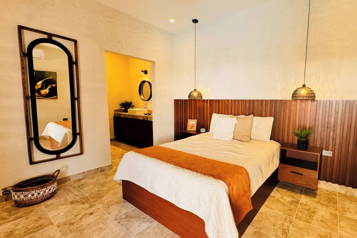 Een moderne slaapkamer met een groot bed, een houten hoofdbord, nachtkastjes, een spiegel en een hangende lamp.