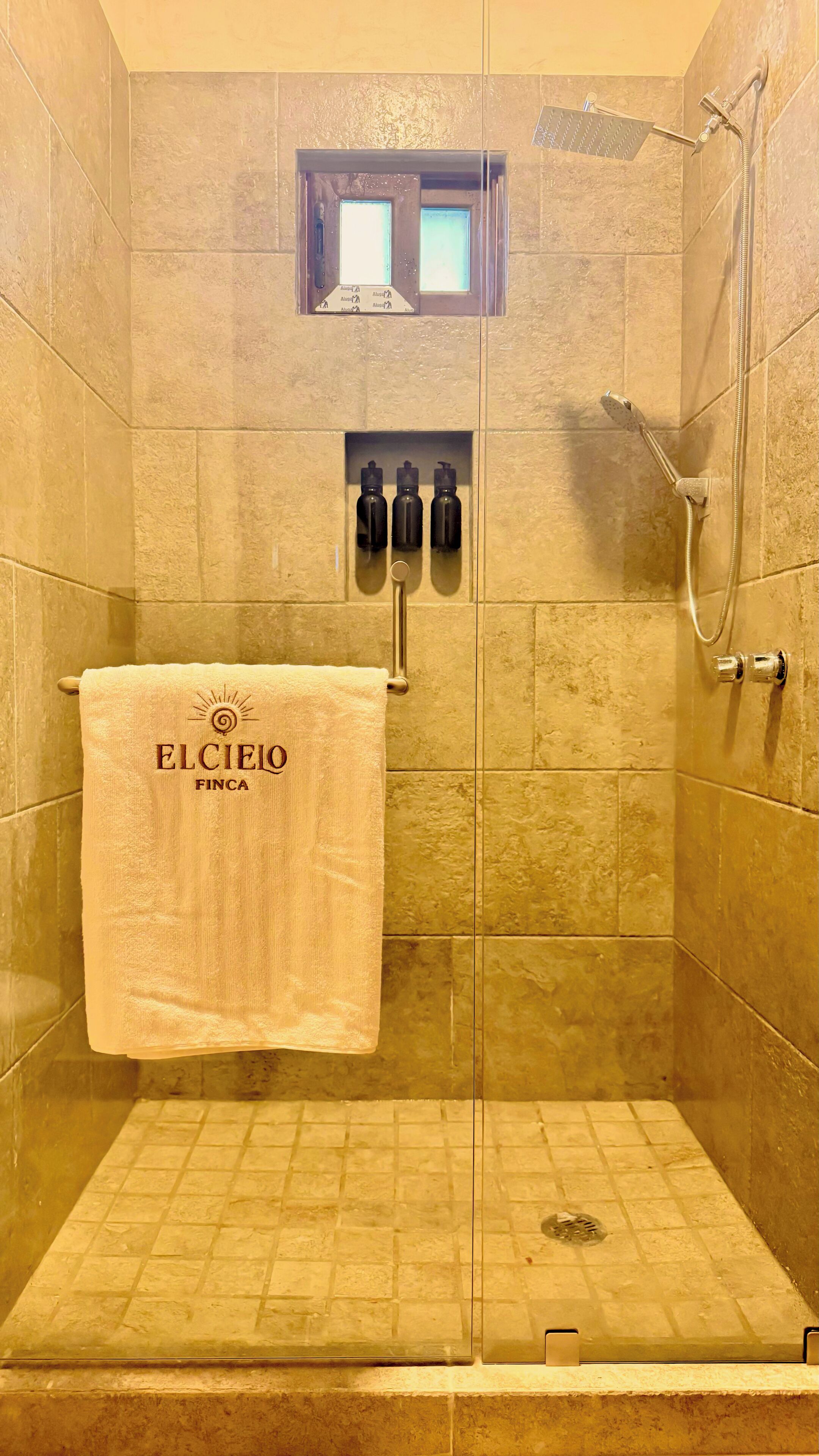 A glass-enclosed shower stall with a beige towel hanging on a rack, a small shelf with bottles, and a showerhead.