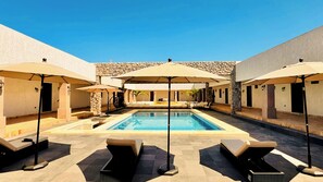 Zona de piscina amb gandules i ombrel·les, parets de pedra i un cel blau clar.