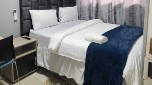Un dormitorio con cabecera capitonada, ropa de cama blanca, manta azul, mesita de noche con lámpara y alfombra estampada.