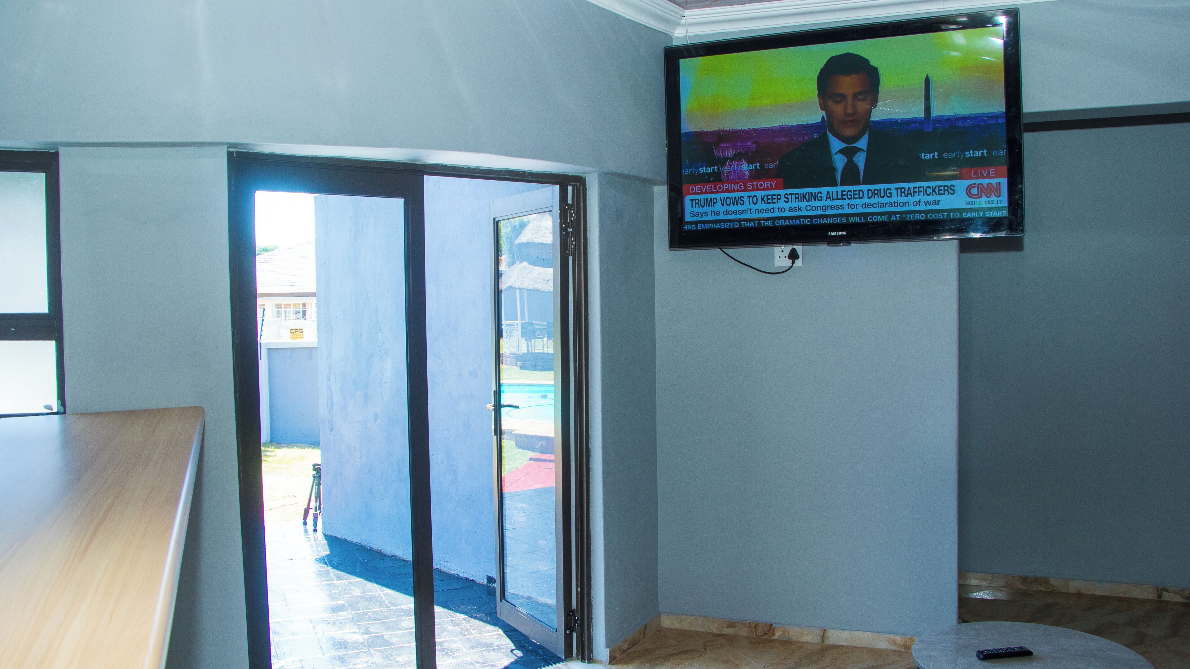 Een kamer met een aan de muur bevestigde televisie waarop een nieuwskanaal wordt vertoond, een glazen schuifdeur naar een buitenruimte en een gedeeltelijk zicht op een houten tafel.