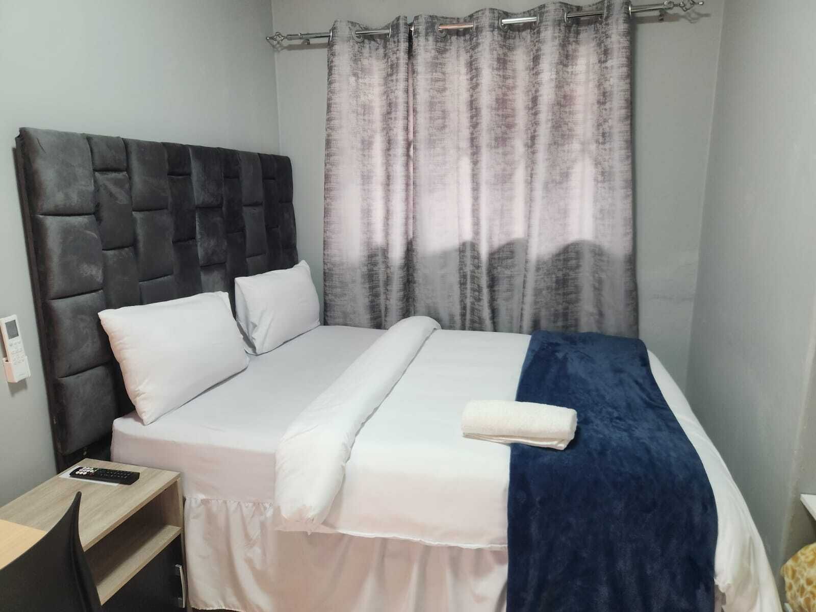 A neatly made bed with white linens and a blue blanket, a dark headboard, and a curtain with a subtle pattern.