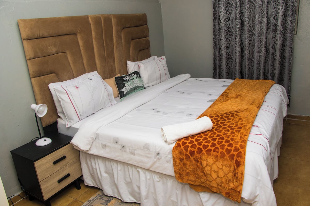 A neatly made bed with a headboard, a bedside table with a lamp, and a patterned curtain.
