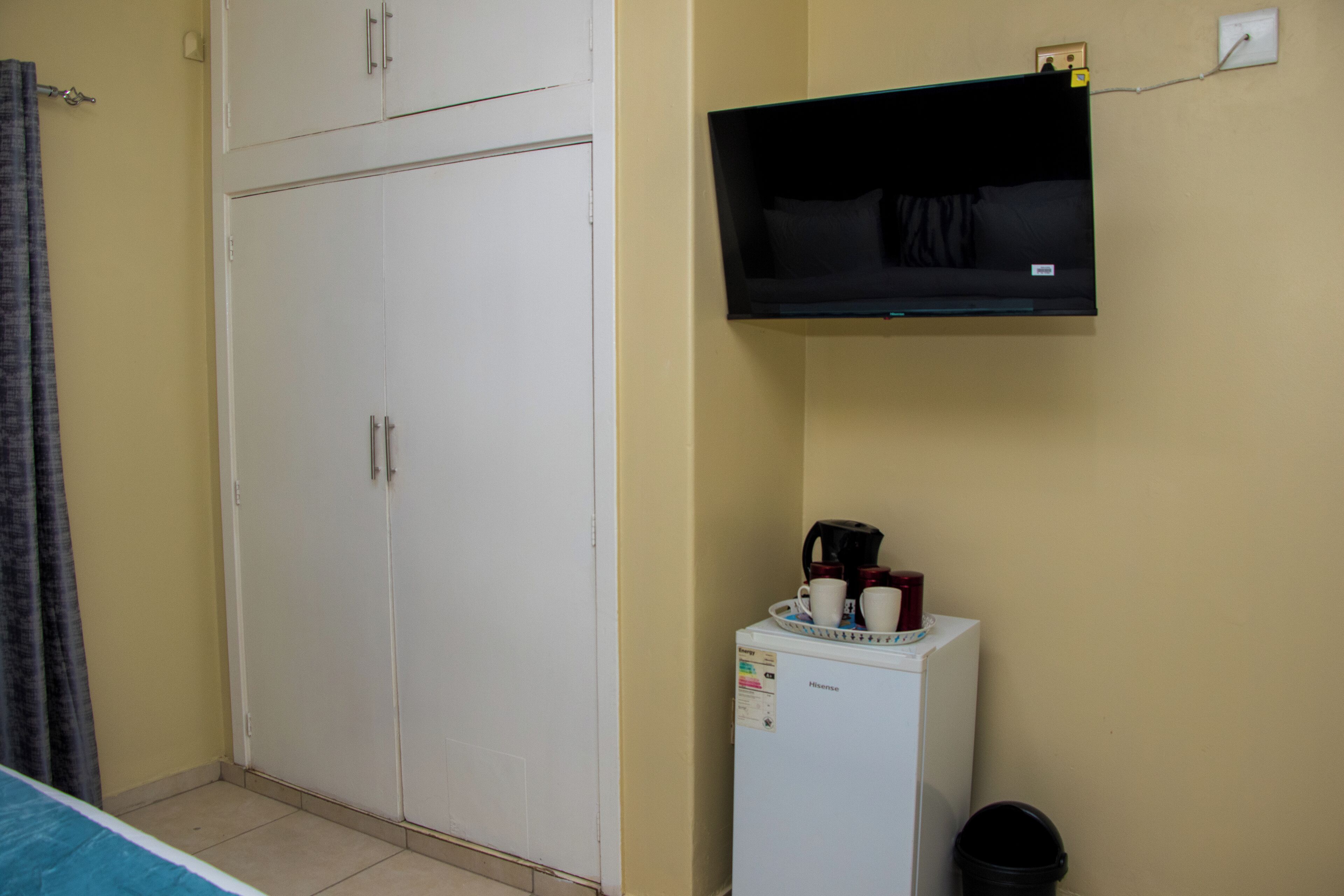 Una habitación con un armario blanco, un pequeño refrigerador blanco con tetera y tazas, y un televisor montado en la pared.