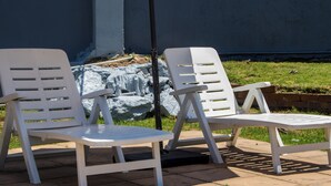 Dos sillones blancos de plástico con una mesita, ubicados en un patio empedrado con un área verde y una pared al fondo.