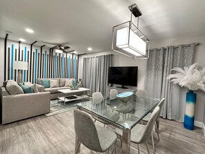 Ein modernes Wohnzimmer mit einem Glastisch, grauen Sofas, einem Flachbildfernseher und einem Deckenventilator.