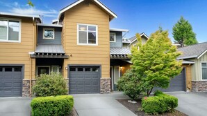 A two-story residential building with a modern design, featuring a garage, a covered porch, and a well-maintained front yard with shrubs and a tree.