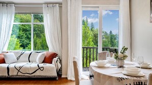 A room with a sofa and a dining table set for two, with a view of trees through large windows.