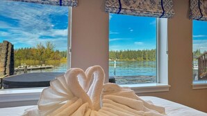 A bed with a heart-shaped towel arrangement, a window with a view of a lake and forest, and a wooden deck outside.