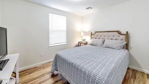 Une chambre à coucher avec un lit, une table de chevet avec une lampe, une fenêtre avec des stores et un téléviseur posé sur un meuble.