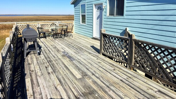 Une terrasse en bois avec un barbecue et des chaises, une maison bleue avec une porte blanche, et une vue sur la plage.