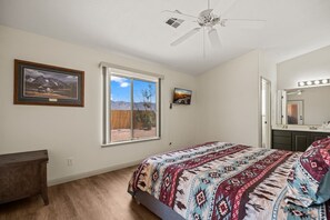 Quarto com cama, ventilador de teto, janela, cômoda e espelho iluminado.