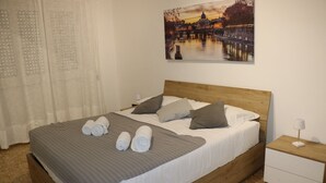 Un letto rifatto con cura, asciugamani arrotolati, una testiera in legno, un comodino con una lampada e un quadro che raffigura una veduta cittadina.