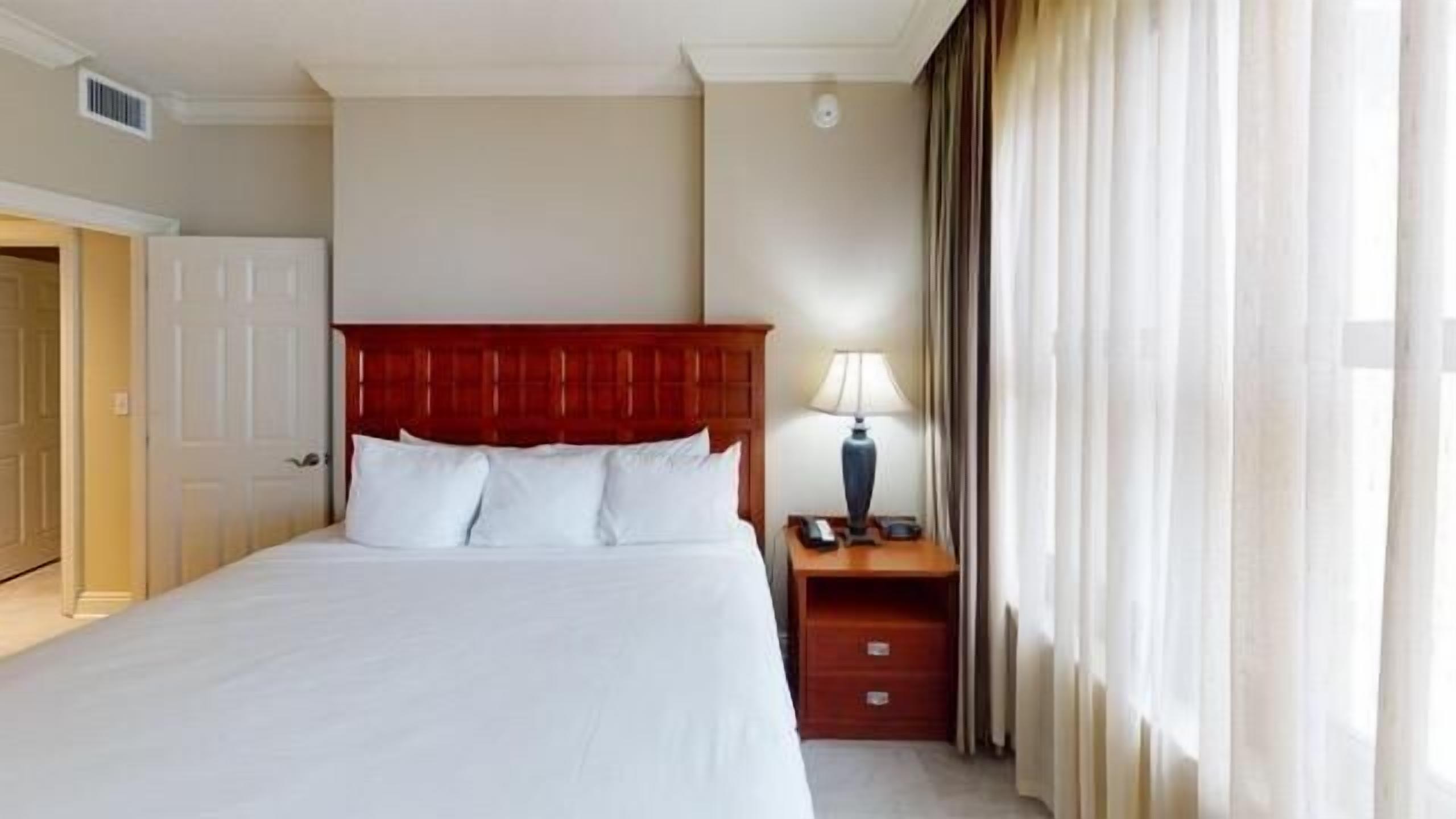 A hotel room with a large bed, a wooden headboard, a nightstand with a lamp, and a window with curtains.