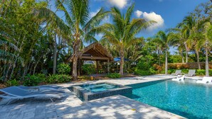 Zona de piscina con sillones reclinables, un jacuzzi y un pabellón rodeado de palmeras y plantas tropicales.