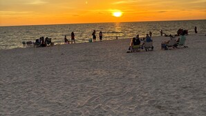 Una playa con gente relajándose y tomando el sol, un atardecer en el fondo y el océano.