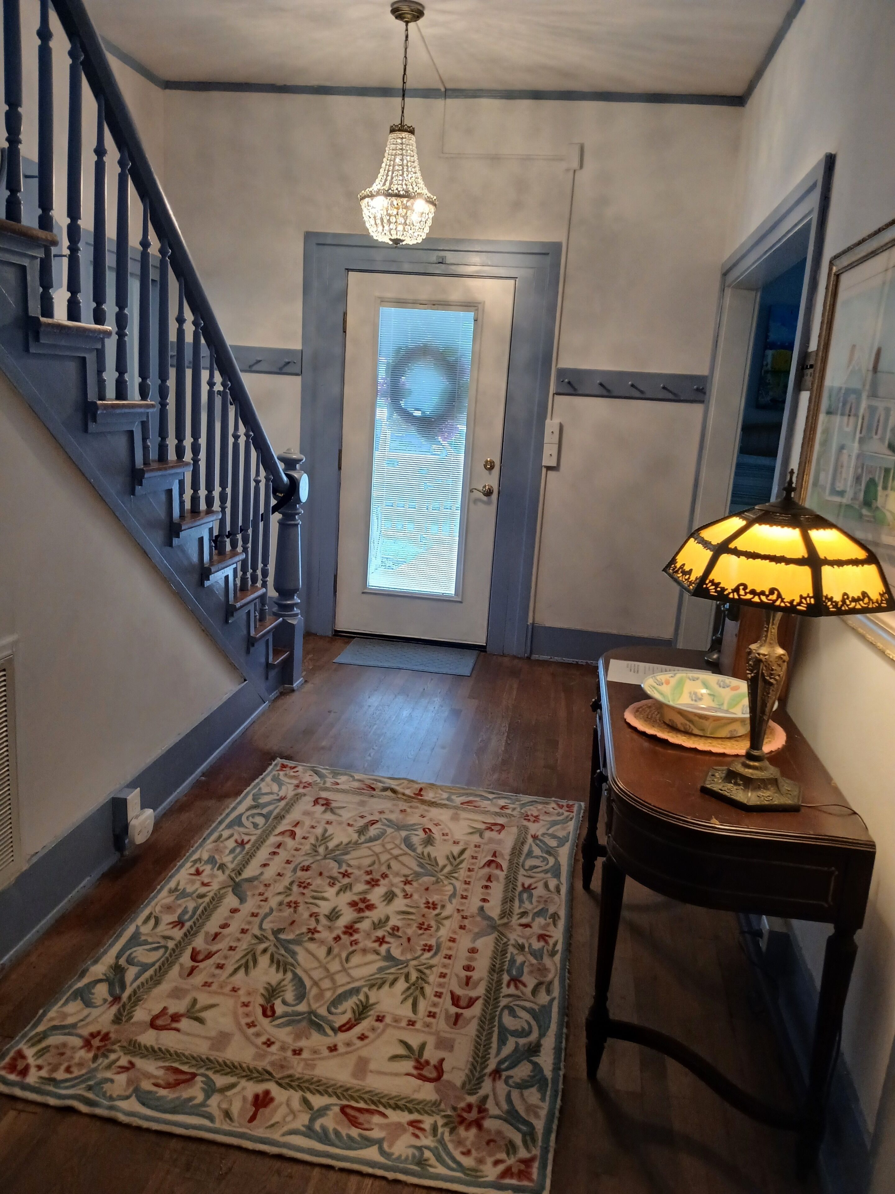A hallway with a wooden staircase, a chandelier, a door with a frosted glass panel, a patterned rug, and a table with a lamp.