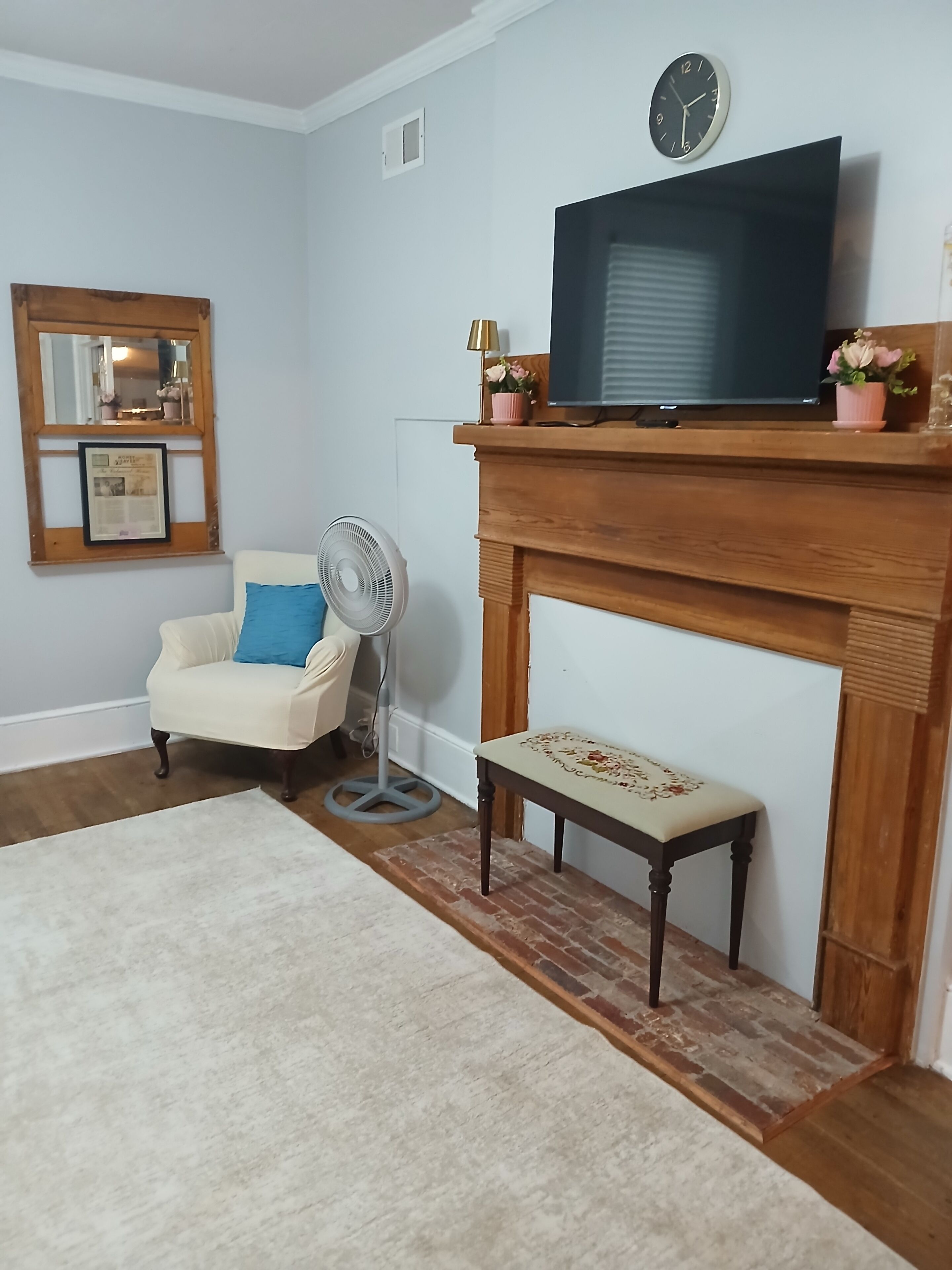 A living room with a television on a wooden mantel, a chair with a cushion, a fan, and a fireplace.