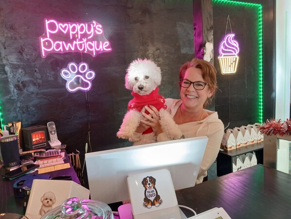 Eine Person hält einen kleinen weißen Hund in einem Raum mit Neonreklamen, auf denen 'Poppy's Pet Boutique' und ein Neon-Eis-Schild zu sehen sind.