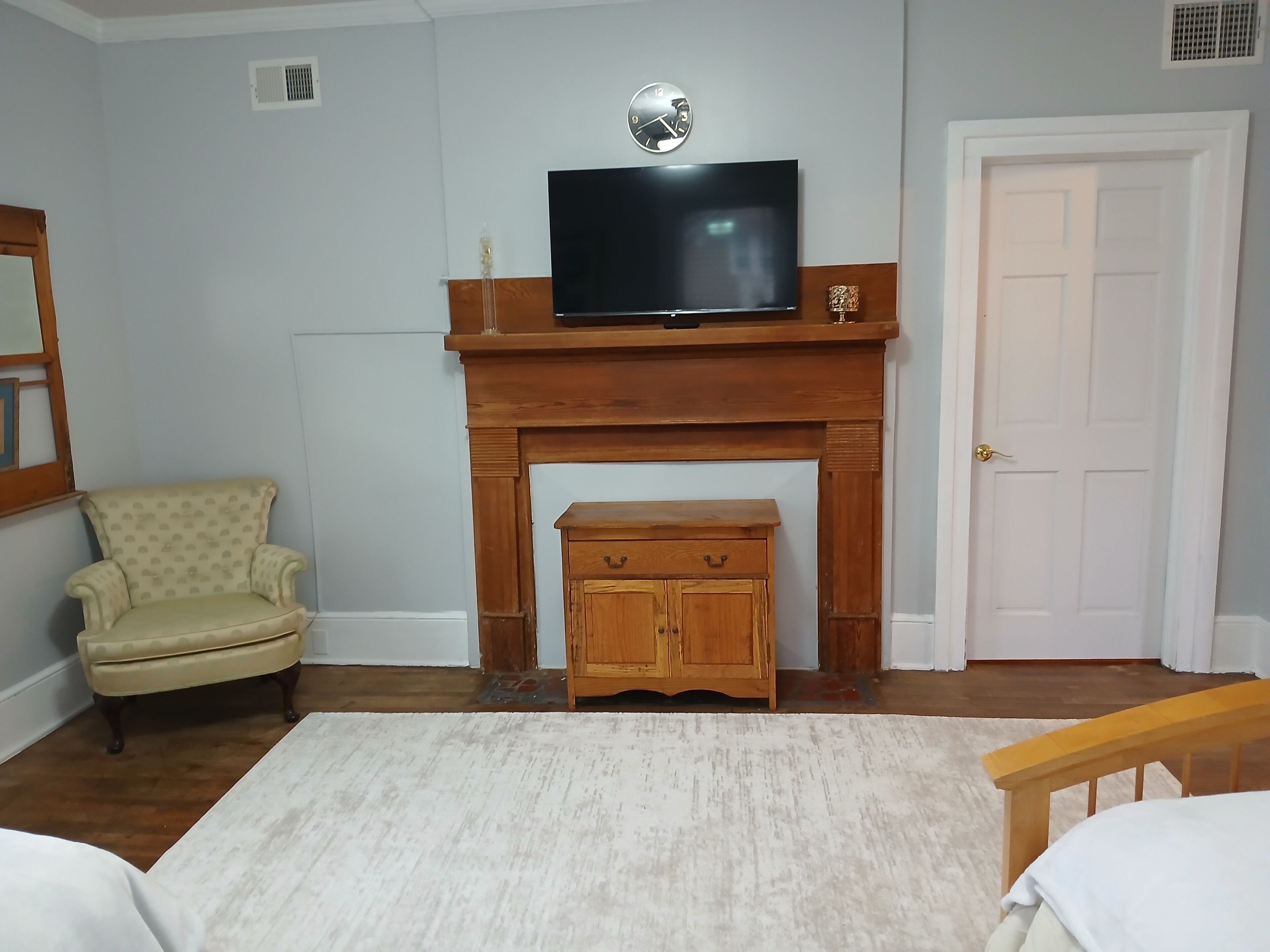 A room with a fireplace, a television mounted above it, a small wooden cabinet below, a patterned armchair, and a white door.