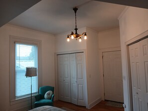 Una habitación con candelabro, ventana con persianas, un sillón azul aguamarina y un conjunto de puertas blancas.