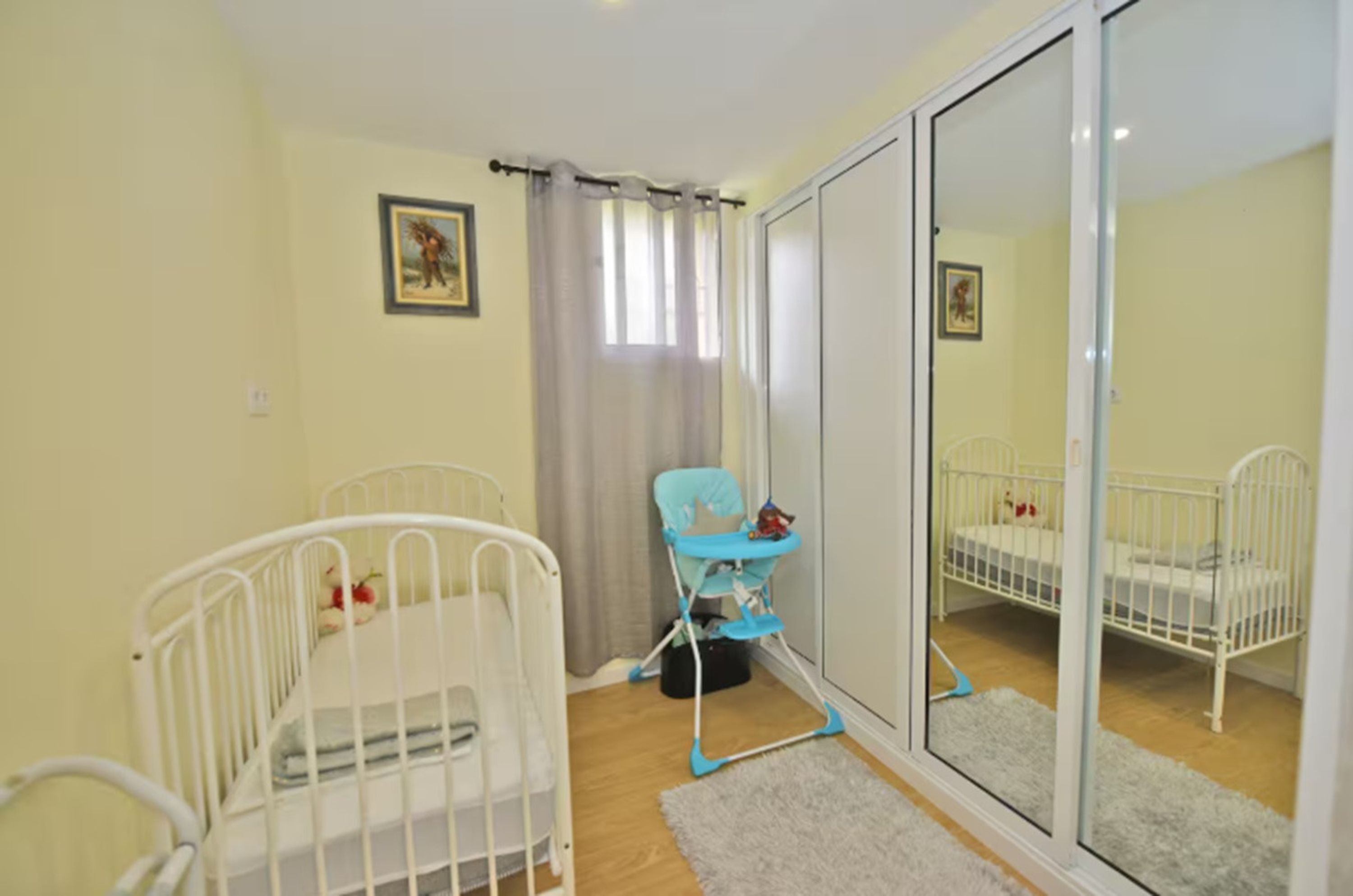 A room with two white cribs, a blue high chair, a sliding glass door, and a small rug.
