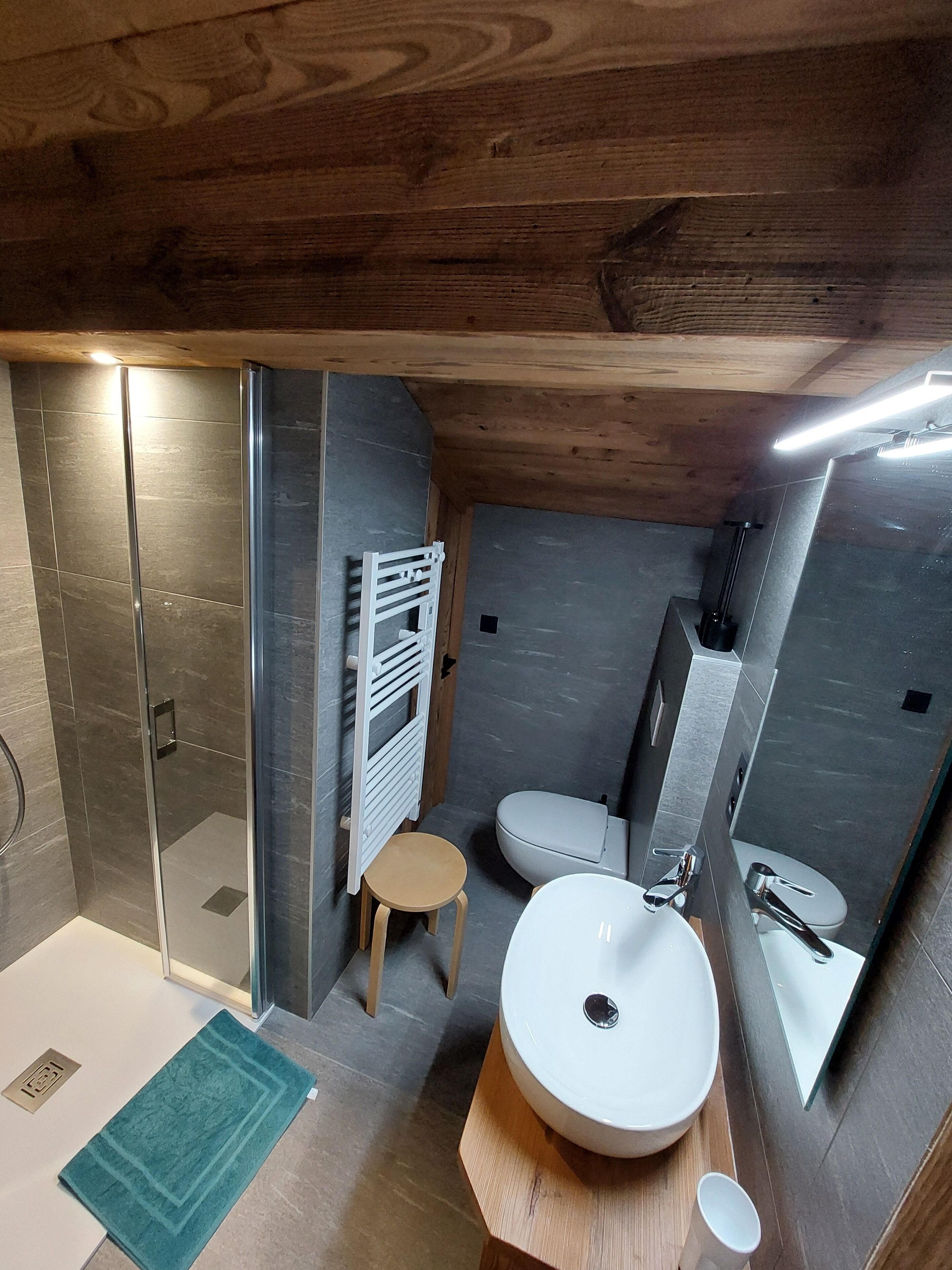 A compact bathroom with a glass shower, a toilet, a sink, and a small stool.