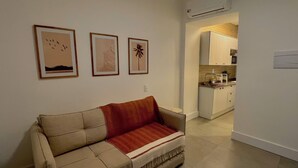 Un salón con un sofá color beige, una manta roja y tres cuadros en la pared. Se ve una cocina al fondo con armarios blancos y un microondas.