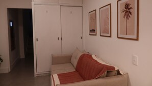 Una habitación con un sofá color beige, una manta roja y blanca, un armario blanco y tres cuadros en la pared.