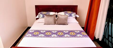 A neatly made bed with a patterned bedspread, a wooden headboard, and a window with curtains.