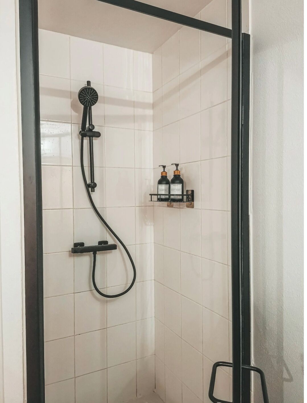 A shower with a handheld showerhead and a handheld hose, a shelf with toiletries, and a black frame around the shower area.