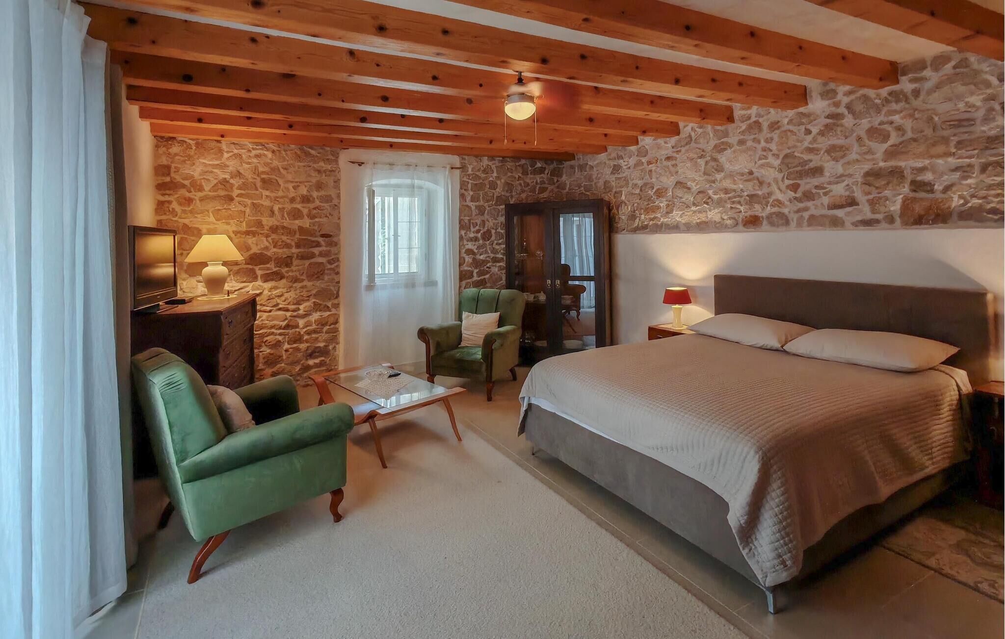 A cozy room with a stone wall, a bed, a green armchair, a wooden ceiling, and a small table with a lamp.