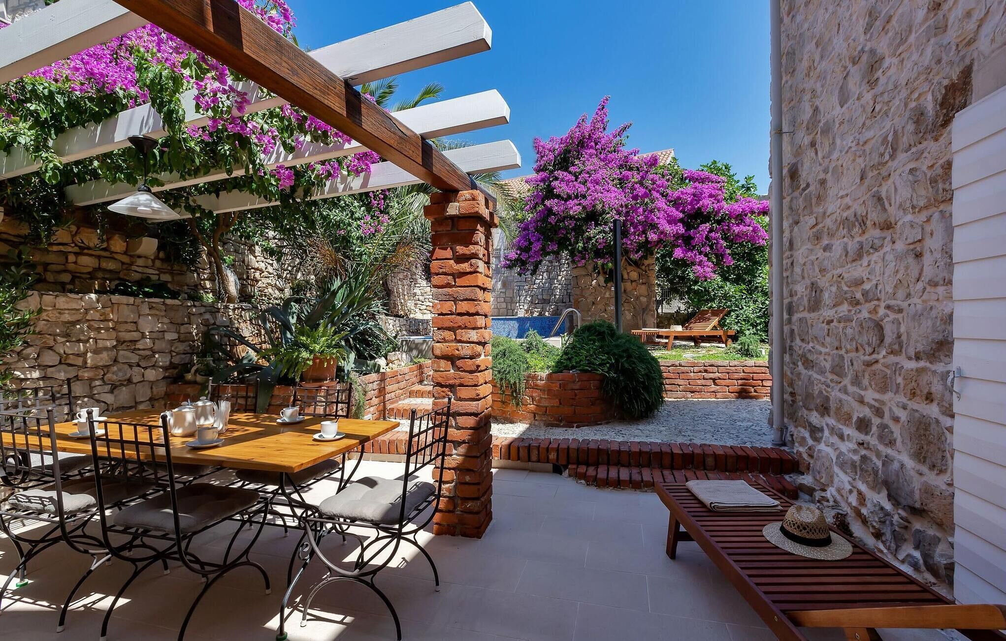 Eine Terrasse im Freien mit einer Steinmauer, einem Holztisch mit Stühlen und einer mit violetten Blumen bewachsenen Pergola.