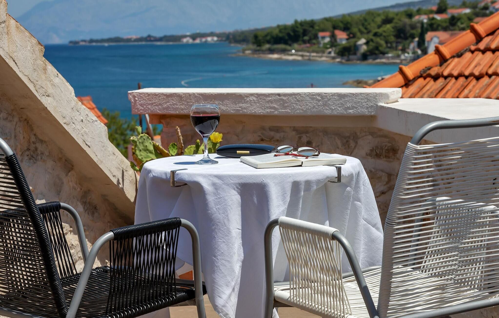 A table with a white tablecloth, a glass of red wine, a book, and glasses on a balcony overlooking a coastal view.