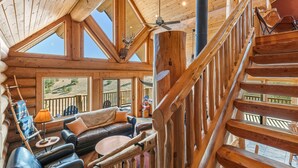 A spacious log cabin interior with a high ceiling, a staircase, a cozy seating area, and large windows offering views of the outdoors. - 45-Acre Florissant Ranch Cabin & Stable (Florissant)
