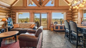 A log cabin interior with a cozy seating area, large windows, and a dining table. - 45-Acre Florissant Ranch Cabin & Stable (Florissant)