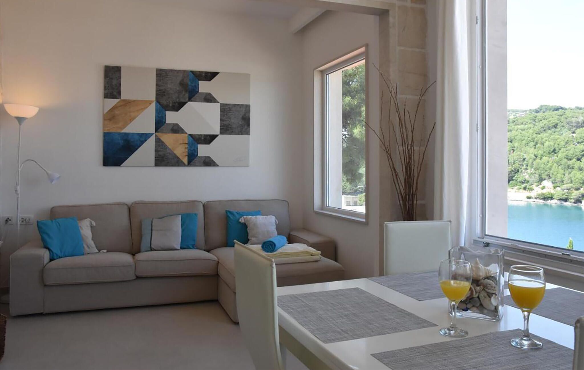 Una sala moderna con un sofá beige, un cuadro abstracto geométrico y vistas a un lago a través de amplios ventanales.
