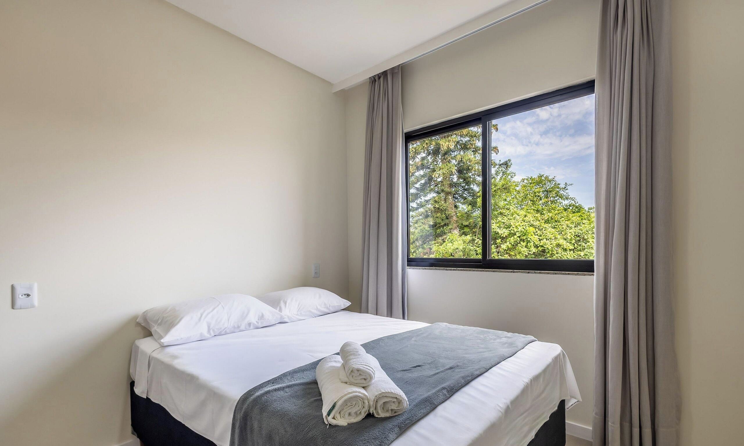 A hotel room with a bed, a window with curtains, and a view of greenery outside.