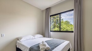 Un lit bien fait, avec du linge de lit blanc et une couverture grise, une fenêtre avec des rideaux et une vue sur les arbres à l’extérieur.