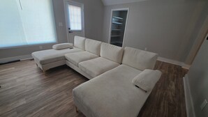 Un sofá modular en una habitación con suelo de madera, una ventana con persianas y una estantería empotrada.