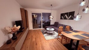 A modern living room with a wooden floor, a sofa, a dining table with chairs, a TV, and a decorative vase.