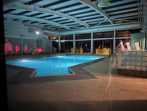 Une piscine intérieure dotée d’un sol carrelé, d’un plafond à structure en forme de grille et d’un espace détente avec des chaises et un mur décoratif.