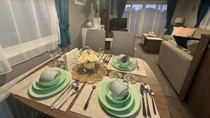 A dining area with a table set for four, including plates, cutlery, and glasses. There is a television in the background and a sofa in the living area. - LuxeHaven - A  Luxury Hot Tub Holiday Home at Tattershall Lakes Country Park (Tattershall)