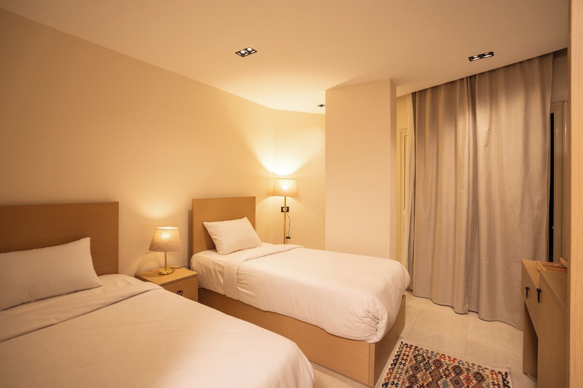 A hotel room with two beds, a nightstand with a lamp, a wooden headboard, a window with curtains, and a small rug.
