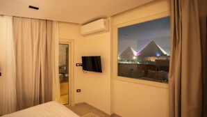 A hotel room with a bed, an air conditioning unit, a TV, and a window with a view of a pyramid-like structure at night. - RIWAQ Pyramids Boutique  (Giza)